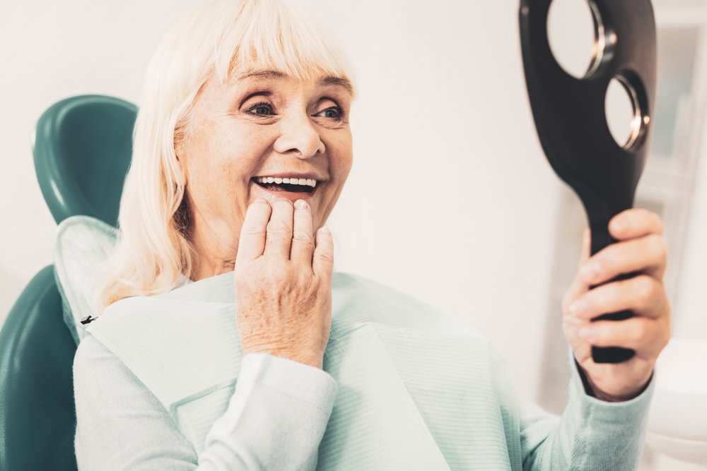 White smile. Close up of mature smiling woman holding mirror and viewing her new denture while touching her chin with hand